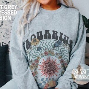 May include: A light grey sweatshirt with a distressed design featuring the word "Aquarius" in a retro font surrounded by flowers, butterflies, and a sun with a psychedelic design. The text "SUNSET DREAMERS.CO" is visible in the bottom right corner.