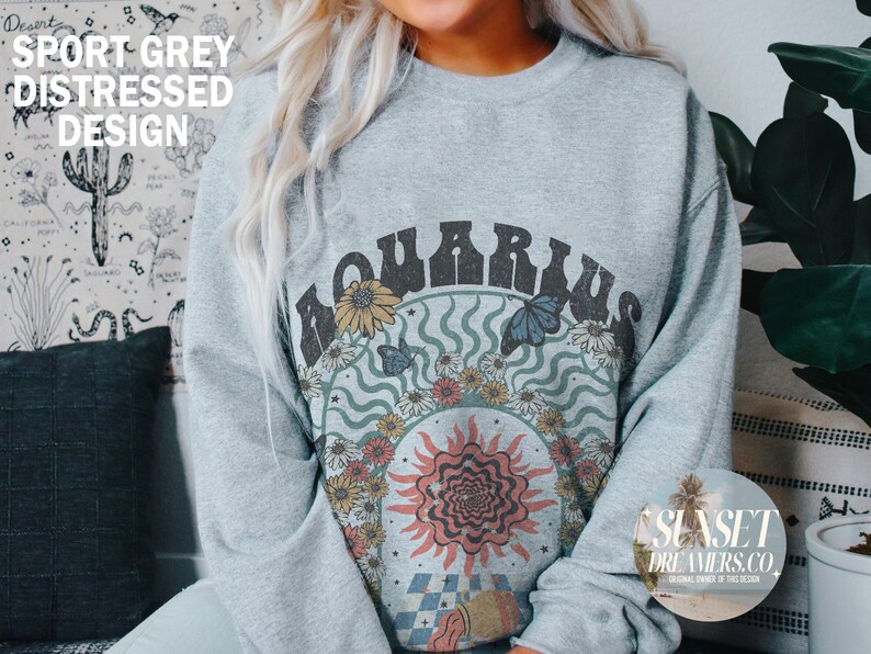 May include: A light grey sweatshirt with a distressed design featuring the word "Aquarius" in a retro font surrounded by flowers, butterflies, and a sun with a psychedelic design. The text "SUNSET DREAMERS.CO" is visible in the bottom right corner.
