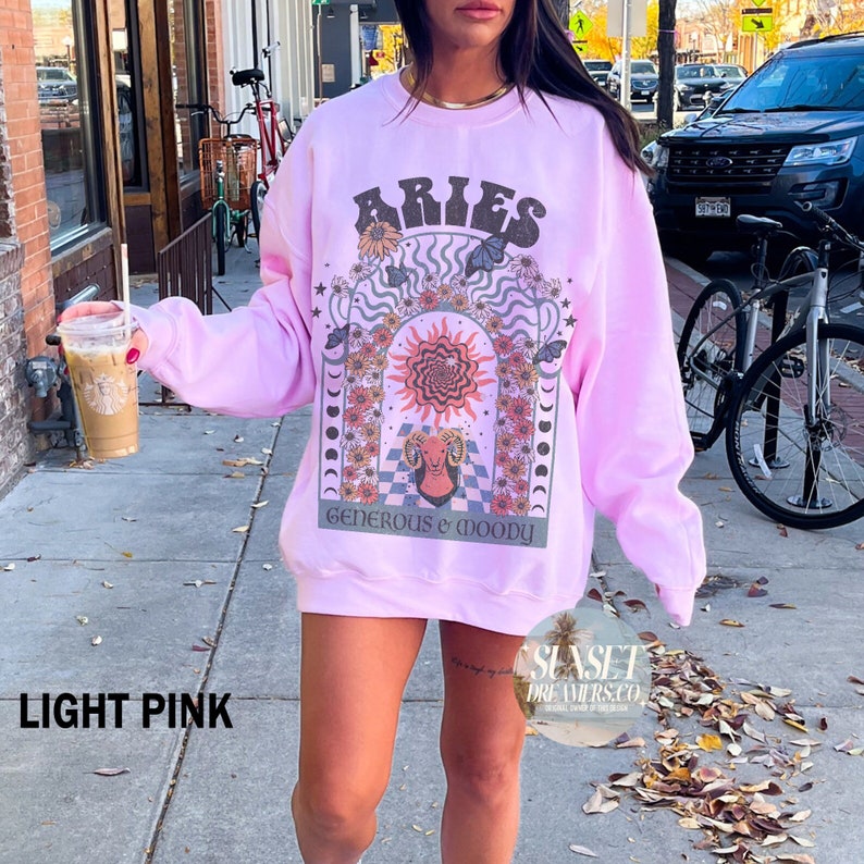Puede incluir: Una sudadera rosa claro con un estampado gr&aacute;fico del signo zodiacal Aries. El gr&aacute;fico presenta un sol, una luna y flores. El texto "Generous & Woody" est&aacute; impreso debajo del gr&aacute;fico.
