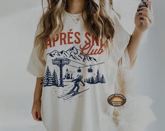 May include: Cream-colored t-shirt with a graphic of a skier, ski lift, and mountains. The text "APRÈS SKI Club" is in red and navy blue. The shirt has a relaxed fit and short sleeves.