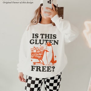 May include: White sweatshirt with a black and orange graphic of a cat pushing a shopping cart full of groceries. The text on the sweatshirt reads "Is this gluten free?"