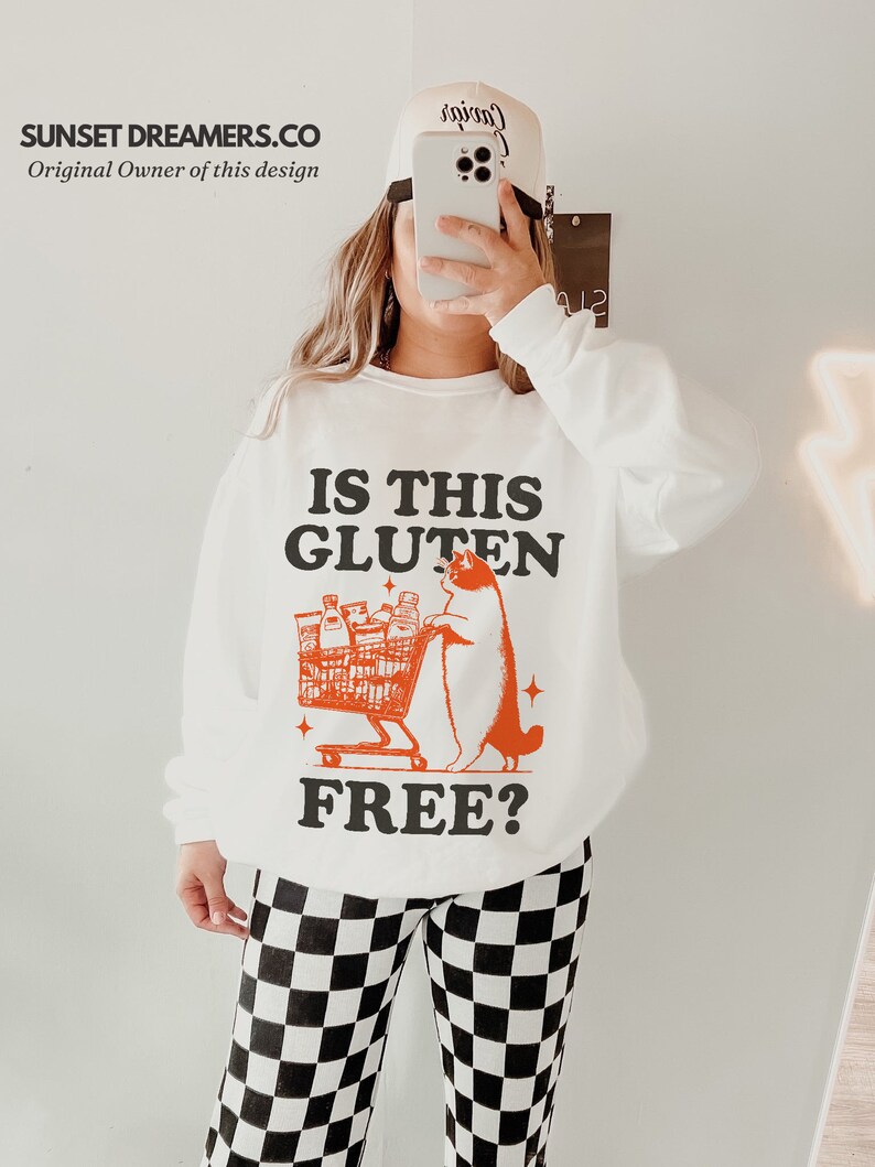May include: White sweatshirt with a black and orange graphic of a cat pushing a shopping cart full of groceries. The text on the sweatshirt reads "Is this gluten free?"