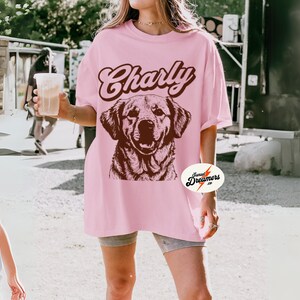 May include: A pink oversized t-shirt with the text "Charly" in brown script and a brown illustration of a dog. The shirt has a small logo on the bottom right corner that says "Sunset Dreamers co."