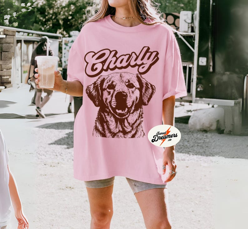 May include: A pink oversized t-shirt with the text "Charly" in brown script and a brown illustration of a dog. The shirt has a small logo on the bottom right corner that says "Sunset Dreamers co."