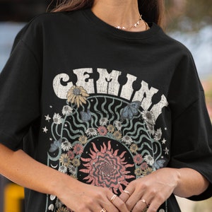 May include: Black t-shirt with a vintage graphic. The design features the word "GEMINI" in distressed white lettering, a sun, flowers, butterflies, and the text "IMAGINATIVE & DEVIOUS".