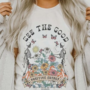 May include: Cream-colored t-shirt with a graphic design featuring two skeleton figures holding hands, surrounded by flowers, butterflies, a sun, and a moon. The text "SEE THE GOOD" and "POSITIVE ENERGY" are displayed above and below the central image.