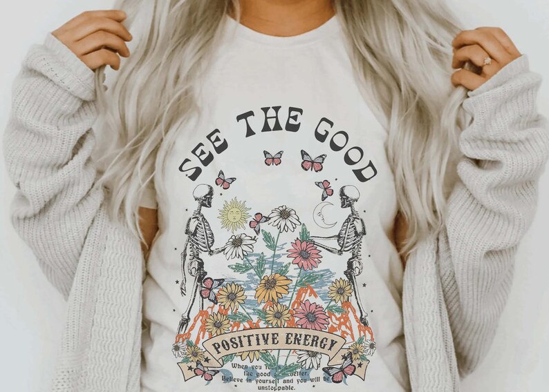 May include: Cream-colored t-shirt with a graphic design featuring two skeleton figures holding hands, surrounded by flowers, butterflies, a sun, and a moon. The text "SEE THE GOOD" and "POSITIVE ENERGY" are displayed above and below the central image.