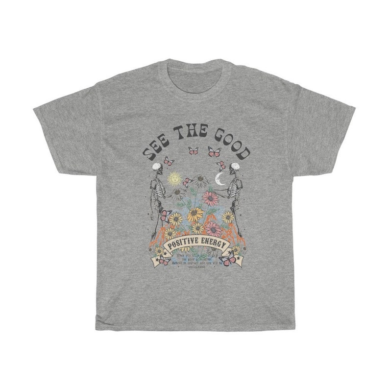 May include: A heather gray t-shirt with a graphic design. The design features two skeletons, flowers, butterflies, and the words "SEE THE GOOD" and "POSITIVE ENERGY". The shirt has a crew neck and short sleeves.
