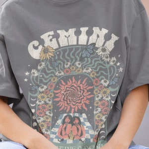 May include: Grey t-shirt with a retro graphic design featuring the word "GEMINI" in a distressed font. The design includes a sun, flowers, butterflies, and two figures. The shirt has short sleeves and a crew neck.