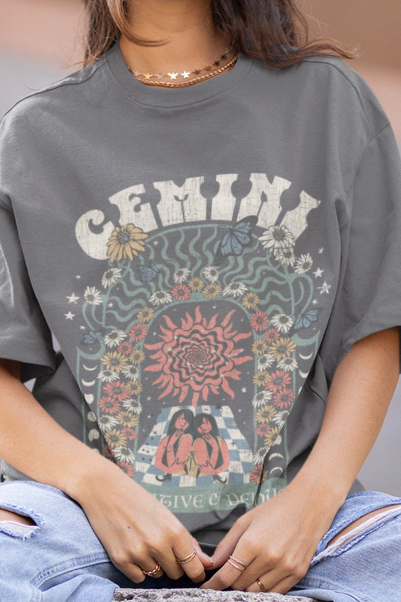 May include: Grey t-shirt with a retro graphic design featuring the word "GEMINI" in a distressed font. The design includes a sun, flowers, butterflies, and two figures. The shirt has short sleeves and a crew neck.