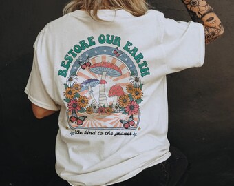 May include: Cream-colored t-shirt with a colorful graphic on the back. The design features mushrooms, flowers, butterflies, and the text "Restore Our Earth" and "Be kind to the planet."