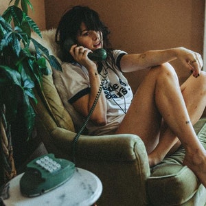 May include: A woman sits in a green armchair, talking on a vintage green rotary phone. She is wearing a white t-shirt with black trim and the text "The Stars" printed on it.