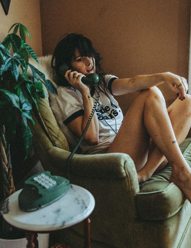 May include: A woman sits in a green armchair, talking on a vintage green rotary phone. She is wearing a white t-shirt with black trim and the text "The Stars" printed on it.