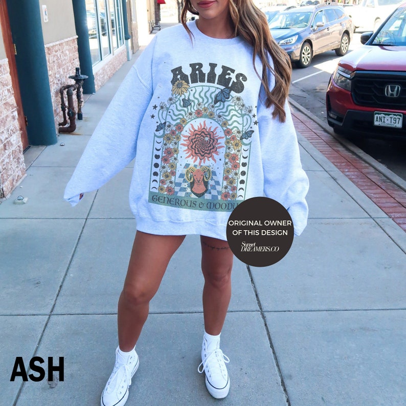 Puede incluir: Una sudadera gris claro con un estampado gr&aacute;fico del signo zodiacal Aries. El gr&aacute;fico presenta un carnero, un sol y flores en un estilo retro colorido. El texto "Aries" est&aacute; en una fuente vintage en negrita. El texto "Generoso y de mal humor" est&aacute; impreso debajo del gr&aacute;fico. El texto "Propietario original de este dise&ntilde;o Sweet Dreamers.co" est&aacute; impreso en un c&iacute;rculo en la esquina inferior derecha de la sudadera.