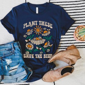 May include: Navy blue t-shirt with a floral design and the text "PLANT THESE SAVE THE BEES". The graphic features daisies, bees, and leaves. The shirt is paired with denim shorts and tan boots.