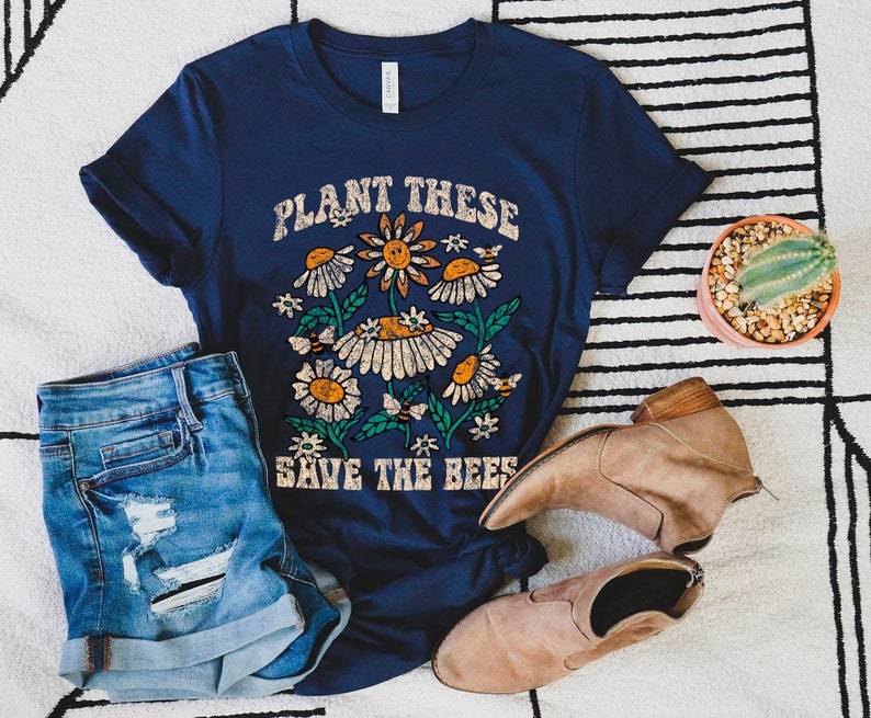 May include: Navy blue t-shirt with a floral design and the text "PLANT THESE SAVE THE BEES". The graphic features daisies, bees, and leaves. The shirt is paired with denim shorts and tan boots.