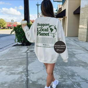 Puede incluir: Una sudadera de color crema con un estampado gr&aacute;fico verde que dice "Support Your Local Planet" con un globo terr&aacute;queo verde y un cactus. El texto debajo del gr&aacute;fico dice "El planeta Tierra es uno de los planetas m&aacute;s &uacute;nicos de nuestro sistema solar. Tiene toda la naturaleza salvaje para superar los eventos adversos y sostener algunas de las formas de vida &uacute;nicas m&aacute;s raras que se conocen. Aunque la Tierra recibe la mayor atenci&oacute;n en nuestro planeta, tambi&eacute;n es la m&aacute;s hermosa y &uacute;nica. Como muchas personas saben, solo mantenemos esta hermosa roca. Haz lo que puedas para ayudar a nuestro planeta. Cuida lo que tienes. Cuida lo que necesitas y disfruta de lo que muchos dan por sentado. Toma solo lo que necesitas y disfruta de lo que muchos dan por sentado. Abraza un futuro sostenible. Propietario original de este dise&ntilde;o: Sweet Dreamer Co."