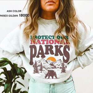 May include: Ash colored sweatshirt with the text "Protect Our National Parks" in a retro font. The graphic features a bear, mountains, and a sunset.