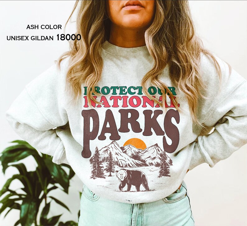May include: Ash colored sweatshirt with the text "Protect Our National Parks" in a retro font. The graphic features a bear, mountains, and a sunset.