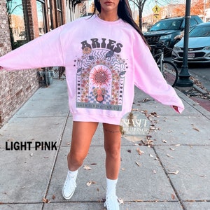 Puede incluir: Sudadera rosa claro con un estampado gr&aacute;fico de un signo zodiacal Aries rodeado de flores y mariposas. El texto "Aries Generous & Moody" est&aacute; impreso debajo del gr&aacute;fico.