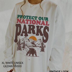 May include: White unisex sweatshirt with a vintage look design. The sweatshirt features a graphic of mountains, a bear, and a sunset with the text "PROTECT OUR NATIONAL PARKS".