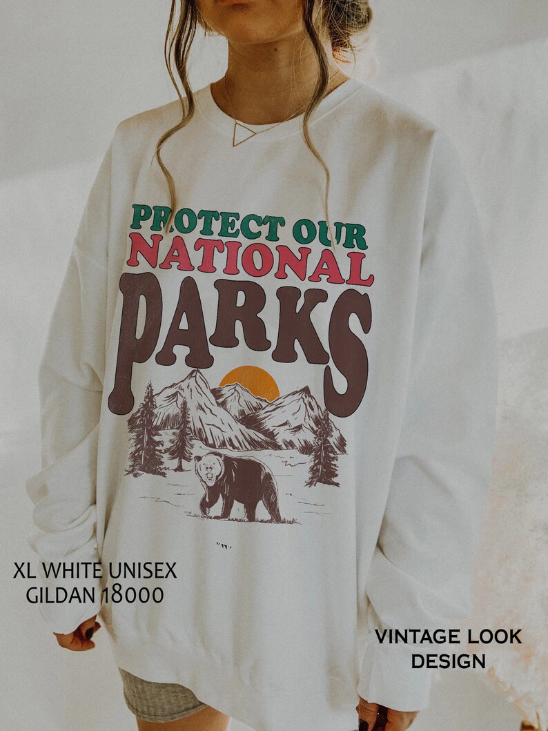 May include: White unisex sweatshirt with a vintage look design. The sweatshirt features a graphic of mountains, a bear, and a sunset with the text "PROTECT OUR NATIONAL PARKS".