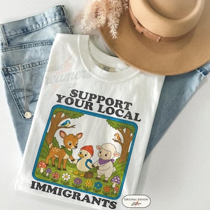 May include: White t-shirt with the text "SUPPORT YOUR LOCAL IMMIGRANTS" and a cartoon illustration of a deer, duck, and sheep holding hands. The shirt is paired with blue jeans and a tan hat.
