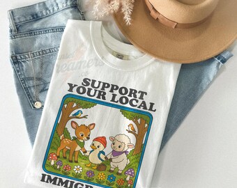 Peut inclure: T-shirt blanc avec l'inscription "SUPPORT YOUR LOCAL IMMIGRANTS" et une illustration de dessin animé d'un cerf, d'un canard et d'un mouton. Le t-shirt est présenté avec un jean bleu et un chapeau beige.