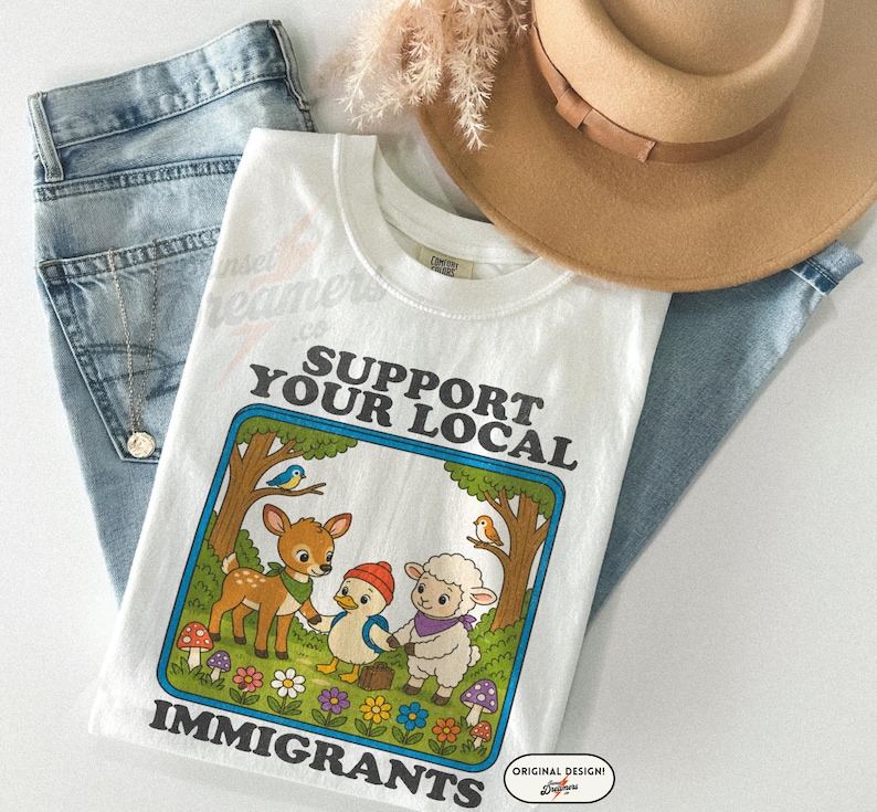 May include: White t-shirt with the text "SUPPORT YOUR LOCAL IMMIGRANTS" and a cartoon illustration of a deer, duck, and sheep holding hands. The shirt is paired with blue jeans and a tan hat.