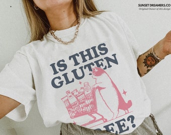May include: White t-shirt with a blue and pink graphic of a cat pushing a shopping cart full of groceries. The text "Is this gluten free?" is printed on the shirt.