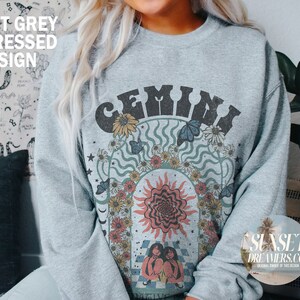 Pode incluir: Uma camisa de moletom cinza com um design gr&aacute;fico que apresenta a palavra "GEMINI" em uma fonte retr&ocirc; cercada por flores, borboletas e um sol. O design &eacute; em tons de azul, verde, amarelo e laranja.