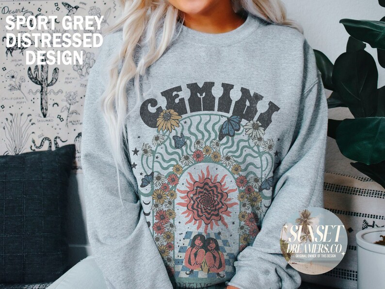 Pode incluir: Uma camisa de moletom cinza com um design gr&aacute;fico que apresenta a palavra "GEMINI" em uma fonte retr&ocirc; cercada por flores, borboletas e um sol. O design &eacute; em tons de azul, verde, amarelo e laranja.