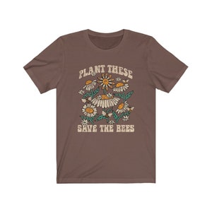 May include: A brown t-shirt with a floral design featuring daisies, bees, and the text "PLANT THESE SAVE THE BEES" in a vintage style. The design includes yellow, white, and green elements.