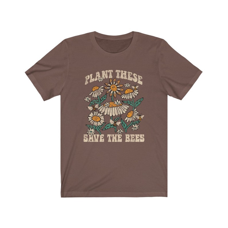 May include: A brown t-shirt with a floral design featuring daisies, bees, and the text "PLANT THESE SAVE THE BEES" in a vintage style. The design includes yellow, white, and green elements.