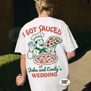 May include: White t-shirt with a graphic of a chef holding a pizza and the text "I got sauced at John and Emily's Wedding".