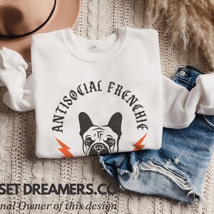 May include: White sweatshirt with a black and white illustration of a French bulldog's head with the text "Antisocial Frenchie" in a black gothic font.