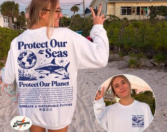 May include: White sweatshirt with a blue graphic print of a shark, a globe, and the text "Protect Our Seas" and "Protect Our Planet". The text also includes the phrase "Embrace a Sustainable Future".
