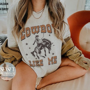 May include: Ash grey unisex Gildan 5000 t-shirt with a vintage-style graphic. The design features the text "COWBOY LIKE ME" in brown lettering, along with an illustration of a cowboy riding a bucking bronco. The shirt also has small star accents.