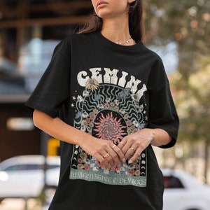 May include: Black t-shirt with a retro graphic. The design features the word "GEMINI" in a distressed font, surrounded by flowers and celestial elements. The design includes a sunburst and the text "IMAGINATIVE & DEVIOUS".