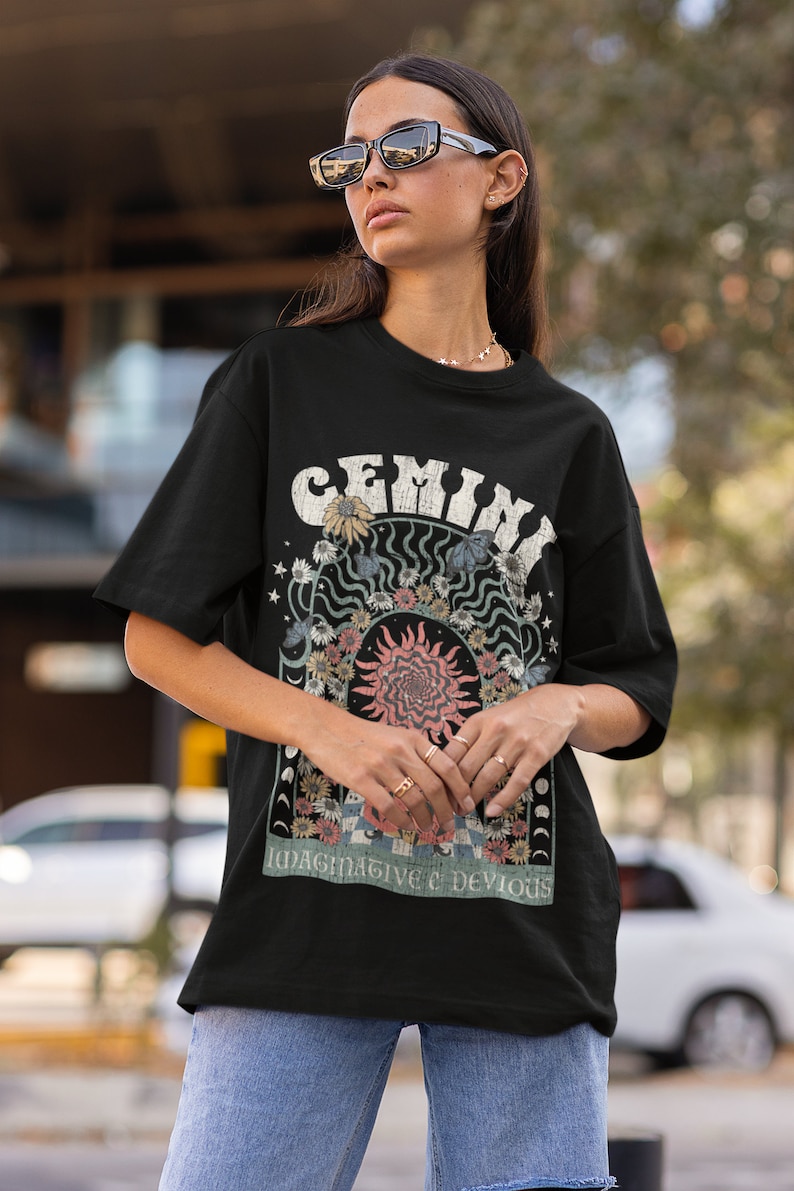 May include: Black t-shirt with a retro graphic. The design features the word "GEMINI" in a distressed font, surrounded by flowers and celestial elements. The design includes a sunburst and the text "IMAGINATIVE & DEVIOUS".