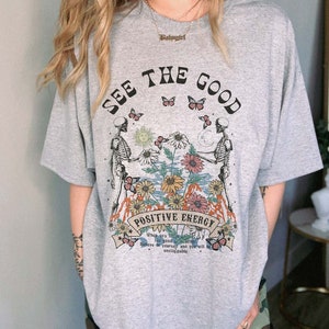 May include: Gray t-shirt with the text "SEE THE GOOD" and "POSITIVE ENERGY". The graphic features skeletons, flowers, butterflies, and the text "When you see the good, believe in yourself and you will be unstoppable."