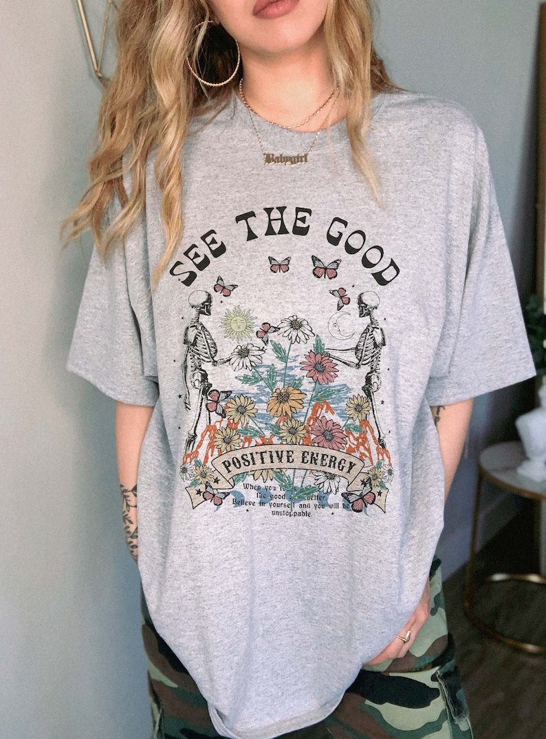 May include: Gray t-shirt with the text "SEE THE GOOD" and "POSITIVE ENERGY". The graphic features skeletons, flowers, butterflies, and the text "When you see the good, believe in yourself and you will be unstoppable."