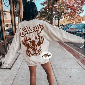 May include: A cream-colored sweatshirt with the word "Charly" in brown text and a brown illustration of a dog on the back. The sweatshirt has a small brown circle with the text "Sunset Dreamers Co." on the bottom left side.