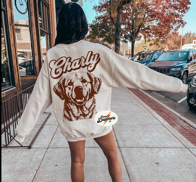 May include: A cream-colored sweatshirt with the word "Charly" in brown text and a brown illustration of a dog on the back. The sweatshirt has a small brown circle with the text "Sunset Dreamers Co." on the bottom left side.