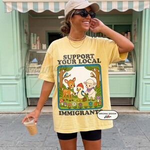 May include: Yellow t-shirt with the text "SUPPORT YOUR LOCAL IMMIGRANT" and a cartoon graphic of a deer, duck, and sheep. The design is framed by trees and flowers. The shirt is worn by a person.