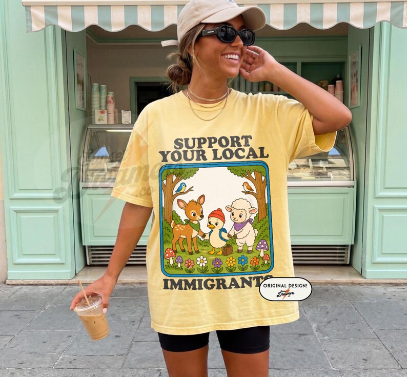 May include: Yellow t-shirt with the text "SUPPORT YOUR LOCAL IMMIGRANT" and a cartoon graphic of a deer, duck, and sheep. The design is framed by trees and flowers. The shirt is worn by a person.