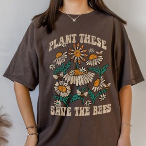 May include: A dark brown t-shirt with a floral design. It features daisies, bees, and the text "PLANT THESE SAVE THE BEES" in a vintage font. The flowers have yellow centres and white petals, with green leaves.