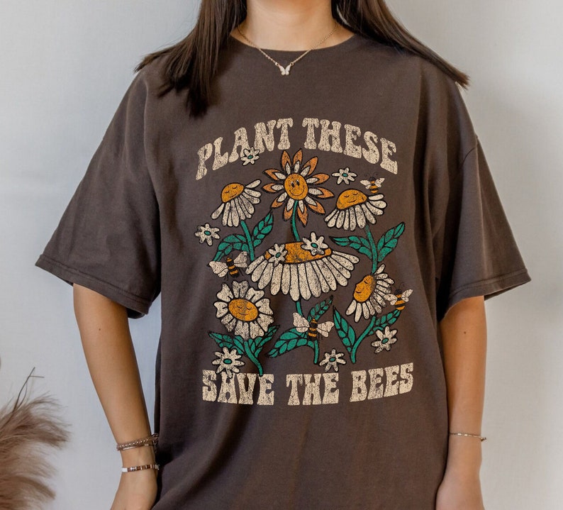 May include: A dark brown t-shirt with a floral design. It features daisies, bees, and the text "PLANT THESE SAVE THE BEES" in a vintage font. The flowers have yellow centres and white petals, with green leaves.