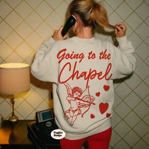 May include: Cream-colored sweatshirt with the text "Going to the Chapel" in red script. The back features a red cupid graphic with a bow and arrow, and red heart accents. The person is wearing red pants.