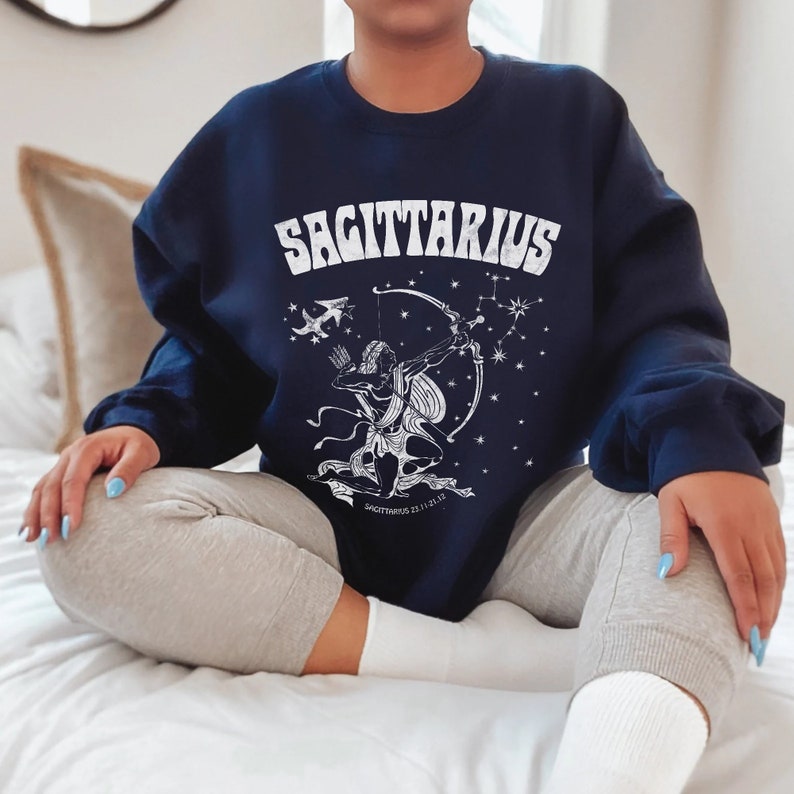 May include: A navy blue sweatshirt with the word "SAGITTARIUS" in white. Below is a white graphic of a centaur with a bow and arrow, constellations, and Sagittarius dates. The sweatshirt is worn with grey sweatpants.